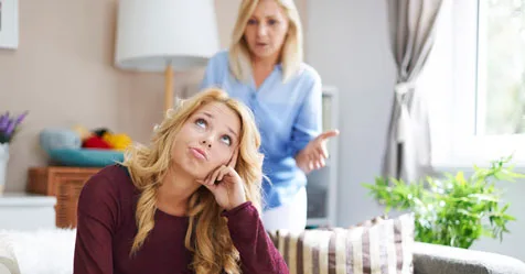 Teen rolling eyes while sitting on a sofa, with a parent talking animatedly in the background.