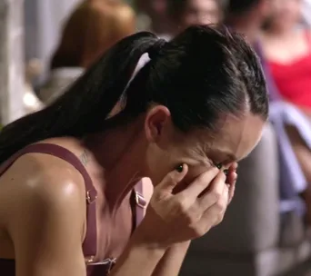 Woman with dark hair in a ponytail crying and covering her face with her hands in a crowded setting.