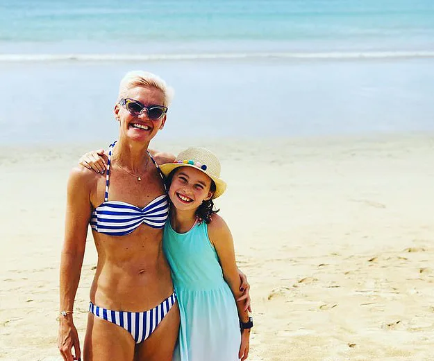 Mother and daughter smiling on a sunny beach; mother in striped bikini, daughter in blue dress with hat.
