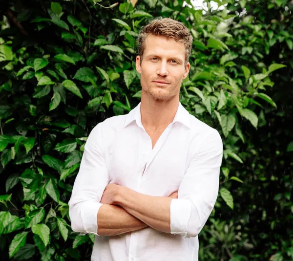 Man in a white shirt stands confidently with arms crossed, surrounded by lush green foliage.