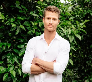 Man in a white shirt stands confidently with arms crossed, surrounded by lush green foliage.