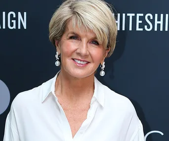 Woman in a white shirt smiling with pearl earrings against a dark background.