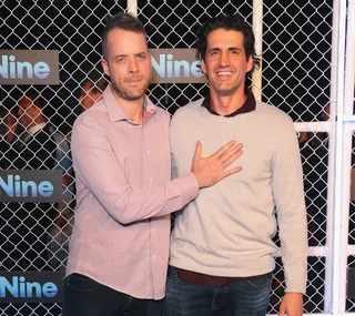 Two men posing in front of a fence with "Nine" logos, smiling at an event.