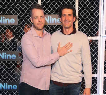 Two men posing in front of a fence with "Nine" logos, smiling at an event.