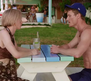 Two people sitting at a colorful picnic table, talking; a drink and a bottle between them, outdoors with a beach bar in background.