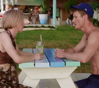 Two people sitting at a colorful picnic table, talking; a drink and a bottle between them, outdoors with a beach bar in background.