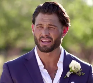 Man in a blue suit with a white rose boutonniere, outdoors in daylight, looks towards the camera, expressing emotion.