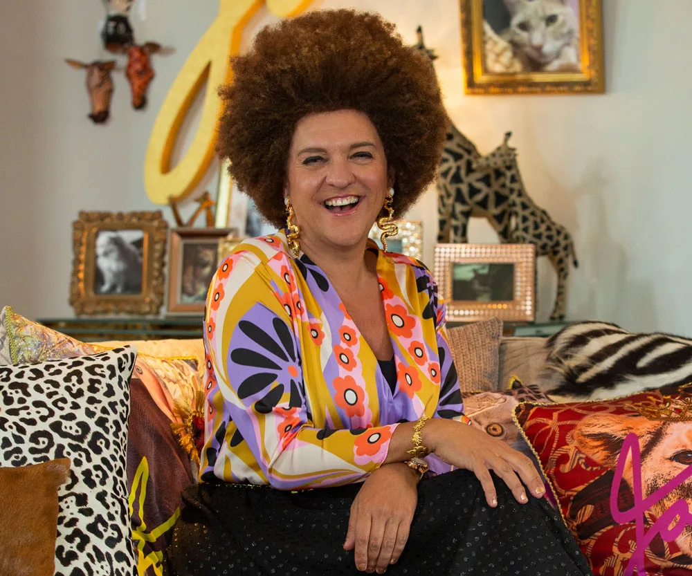 A person with an afro hairstyle smiles while sitting in a vibrant room with eclectic decor and animal print cushions.