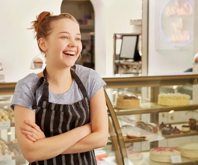 Young woman in striped apron smiling in a bakery with cakes in display.