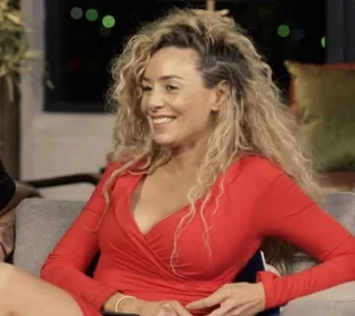 A woman with long curly hair in a red dress sits on a couch, smiling.