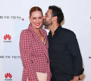 Couple poses on red carpet; woman in pink plaid suit smiles, man in black shirt kisses her cheek.