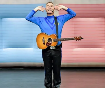 Man in blue shirt with guitar, hands near ears, smiling on stage with red and blue background.