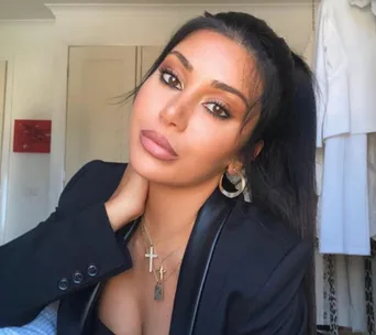 Young woman with long dark hair, wearing a black blazer and necklaces, poses indoors with a pensive expression.