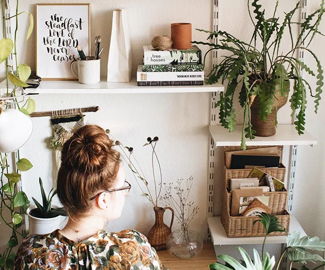 Woman with glasses facing bookshelves with plants, books, and decor in a cozy room.