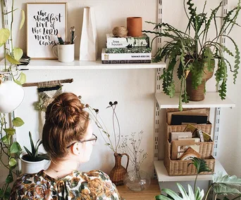 Woman with glasses facing bookshelves with plants, books, and decor in a cozy room.