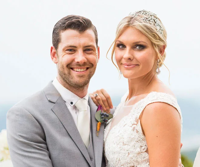 A bride and groom stand smiling, with the groom in a gray suit and the bride in a lace dress and tiara.