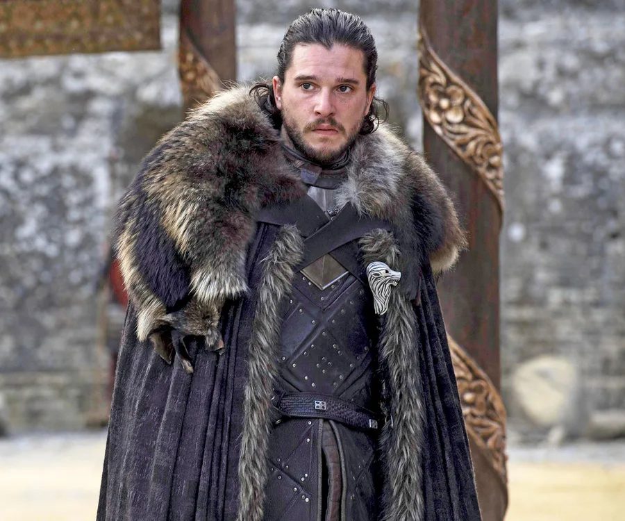 Man in fur cloak and armor stands outdoors, serious, with long dark hair.