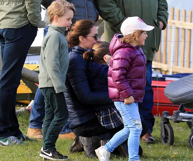 Prince George, Mia Tindall, and a woman sitting on grass during an outdoor event, surrounded by people.