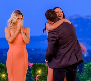 A woman in an orange dress reacts emotionally as a couple hugs joyfully against a scenic outdoor backdrop.