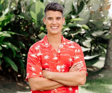 Man in a red Hawaiian shirt smiling with arms crossed, standing in front of lush greenery.