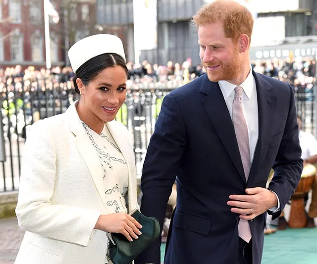 Meghan Markle and Prince Harry smiling outside, Meghan in a white outfit and Harry in a suit.