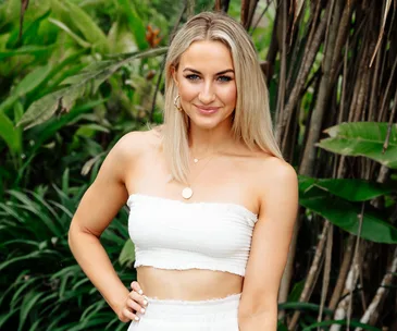 A woman in a white strapless top smiles, standing outdoors with lush greenery in the background.