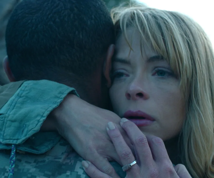 A woman with blond hair hugs a person in military attire, looking pensive and emotional.