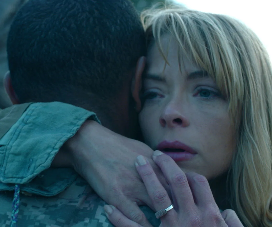 A woman with blond hair hugs a person in military attire, looking pensive and emotional.