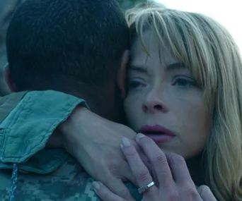 A woman with blond hair hugs a person in military attire, looking pensive and emotional.