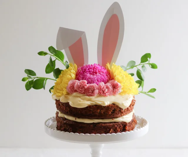 Easter carrot cake with cream cheese frosting, topped with flowers and bunny ears on a white cake stand.