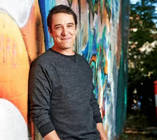 Man in a dark sweater leans against a colorful graffiti wall, smiling warmly at the camera.