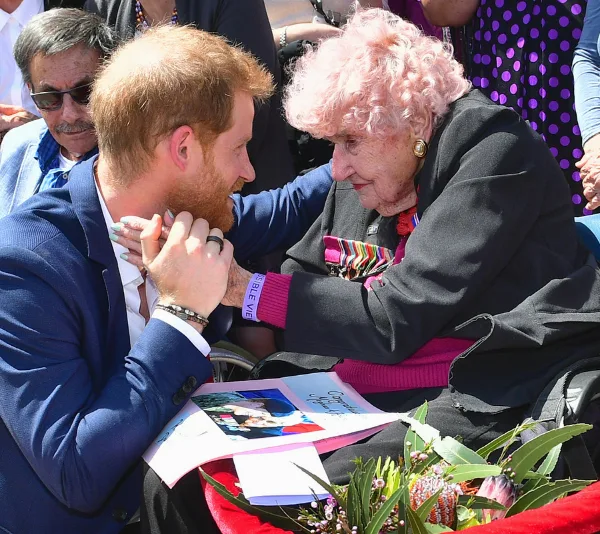 Prince Harry kneels to embrace a smiling elderly woman outdoors, with flowers and a booklet in view.