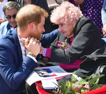 Prince Harry kneels to embrace a smiling elderly woman outdoors, with flowers and a booklet in view.