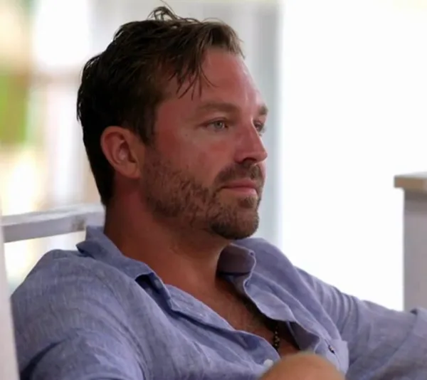 A man in a light shirt is sitting casually, looking pensive, with a blurred background.