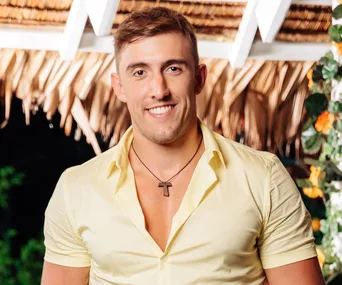 Man in a yellow shirt smiles, standing under a pergola with foliage and flowers in the background.
