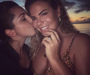 Two women smiling at sunset; one kisses the other's cheek, with an engagement ring on display.