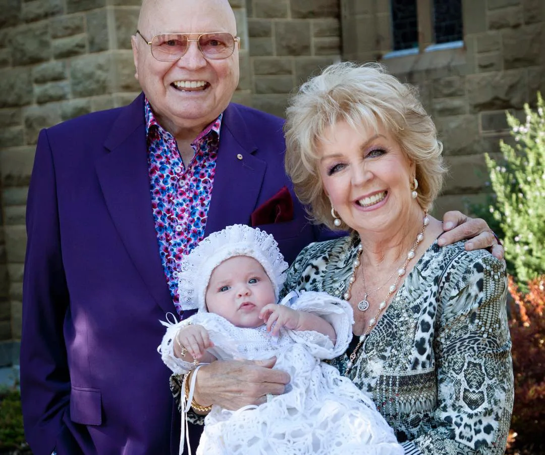 Elderly couple smiling while holding a baby in white christening attire outdoors.