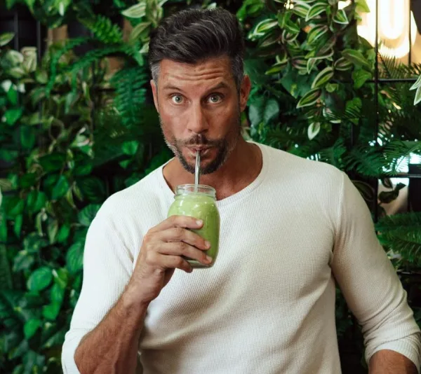 Man sipping green smoothie in front of lush green plants.