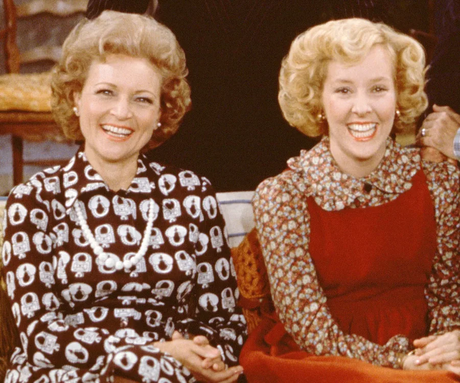 Two women sitting and smiling, wearing patterned dresses, from a classic TV show.