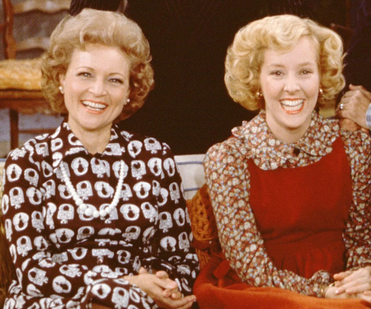 Two women sitting and smiling, wearing patterned dresses, from a classic TV show.