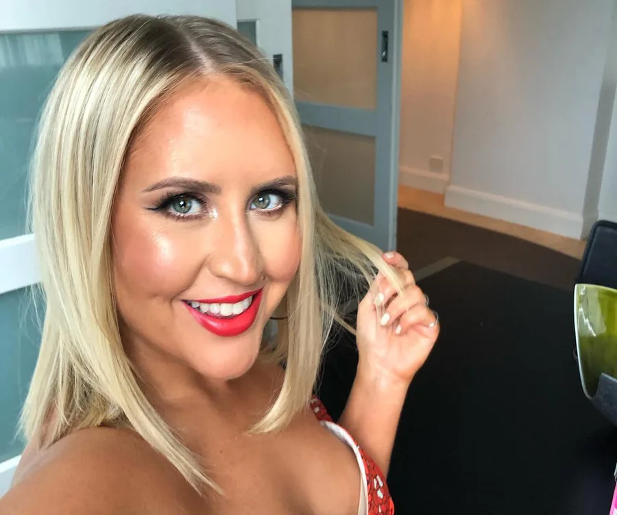 Blonde woman smiling, wearing red lipstick and holding her hair in a modern interior setting.