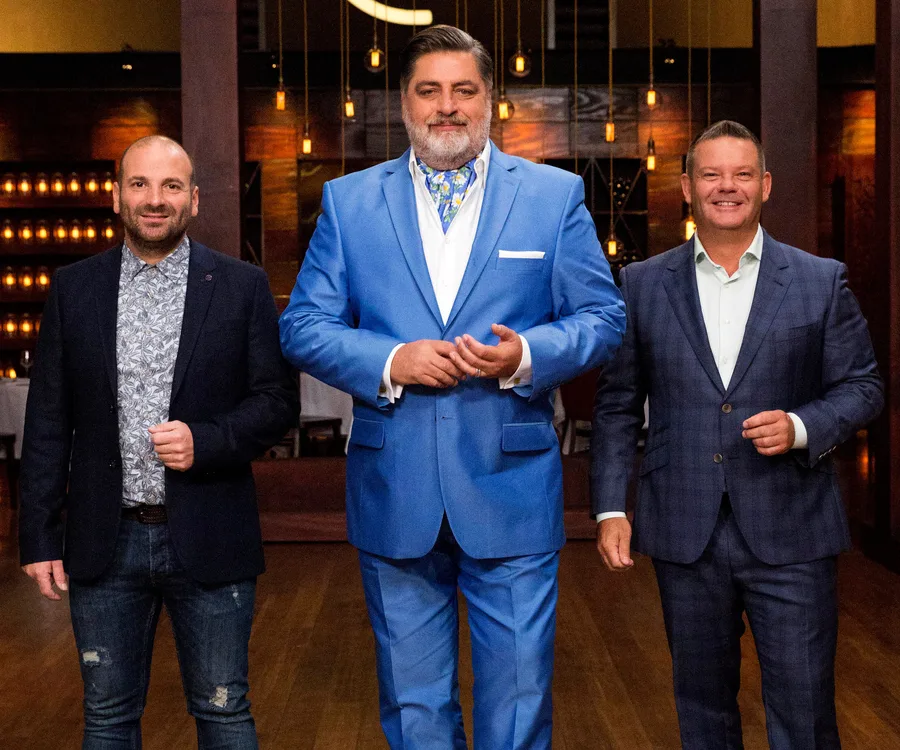 Three men, likely MasterChef Australia judges, stand smiling in a warmly lit room, wearing suits.