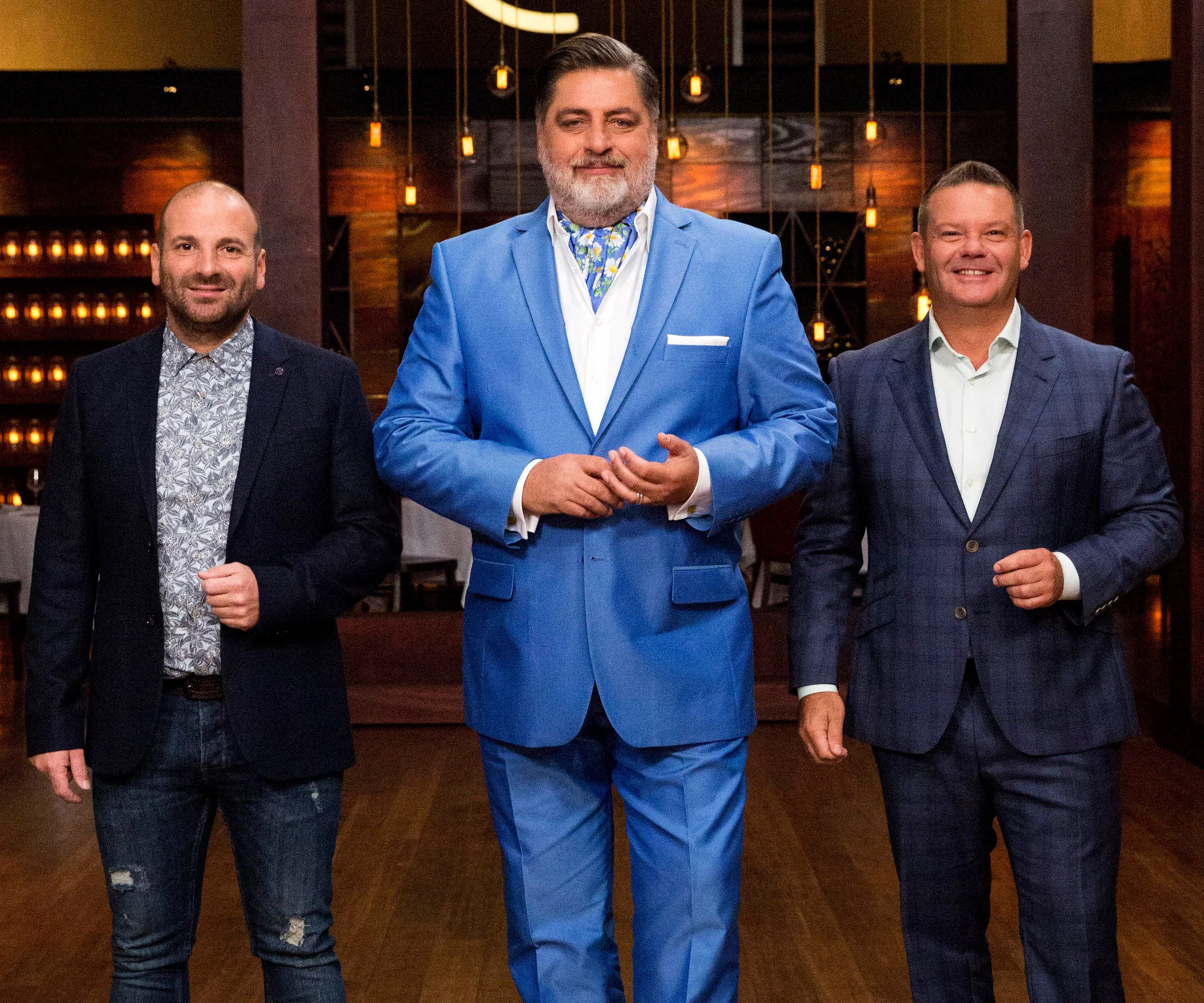 Three men, likely MasterChef Australia judges, stand smiling in a warmly lit room, wearing suits.