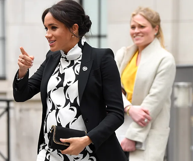 Meghan Markle in a patterned dress and black blazer, smiling and gesturing while holding a black clutch.