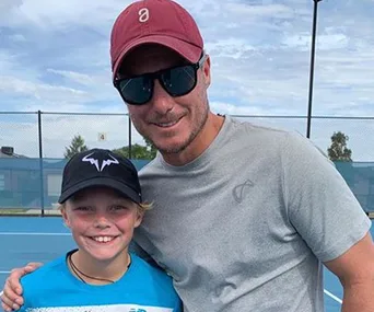 Father and son on a tennis court, dad in sunglasses and cap, son wearing a blue shirt and black cap with a logo.