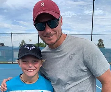 Father and son on a tennis court, dad in sunglasses and cap, son wearing a blue shirt and black cap with a logo.