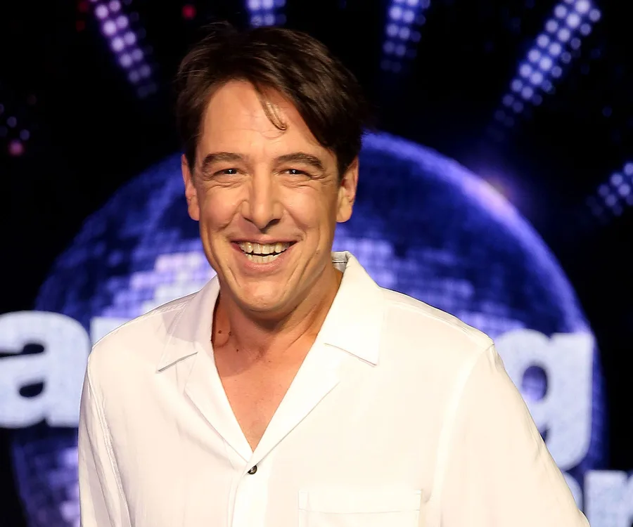 Smiling man in a white shirt, in front of a "Dancing with the Stars" background with a sparkling globe.