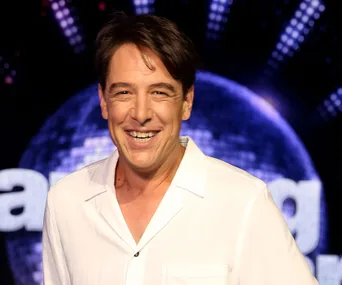 Smiling man in a white shirt, in front of a "Dancing with the Stars" background with a sparkling globe.
