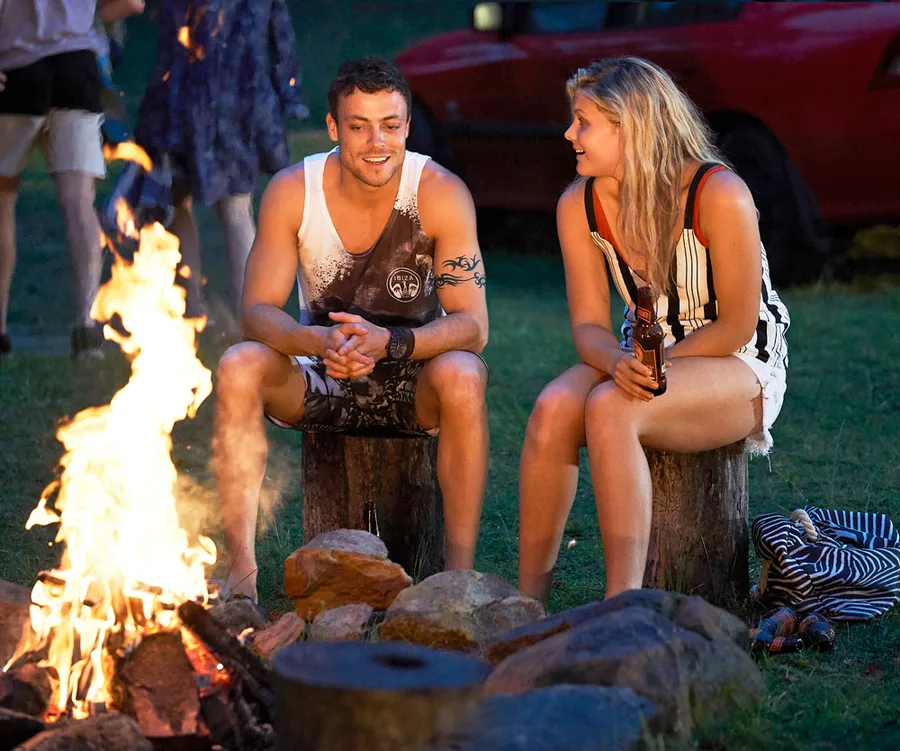 Man and woman sitting by a campfire, smiling and holding drinks.