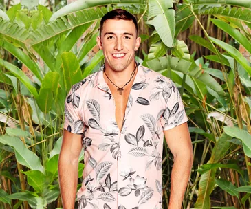 Man smiling in a tropical shirt, standing in front of lush green tropical plants.